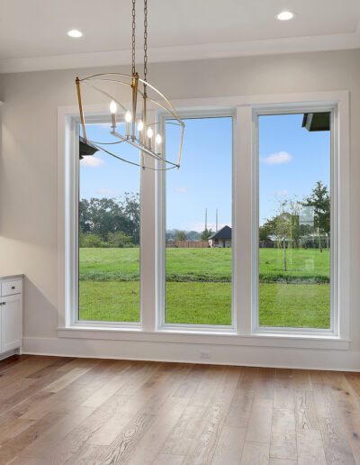 A bright, empty room with large windows overlooking a green yard, featuring white cabinetry and a chandelier.