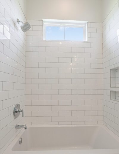 A clean, white-tiled bathroom with a bathtub and a window letting in natural light.