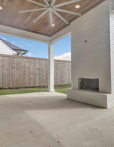 Spacious covered patio with a fireplace and ceiling fan, overlooking a lawn with a wooden fence.